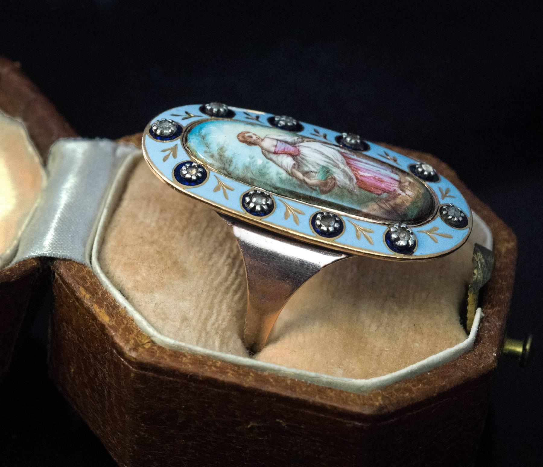 An antique, circa 1790 / 1810, rose 14K gold long ring features a painted enamel pastoral scene (idealized country life) of a peasant girl carrying a basket of greens. The painted enamel miniature is set in a sky blue enamel frame embellished with