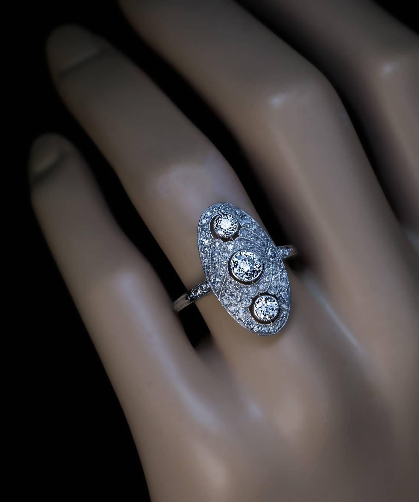 A vintage platinum engagement ring is vertically bezel-set with three bright white old European cut diamonds (approximately 0.34 ct, 0.17 ct, 0.15 ct; F color, VS1, VS2, SI1 clarity) surrounded by ornate oval frame densely set with old single cut