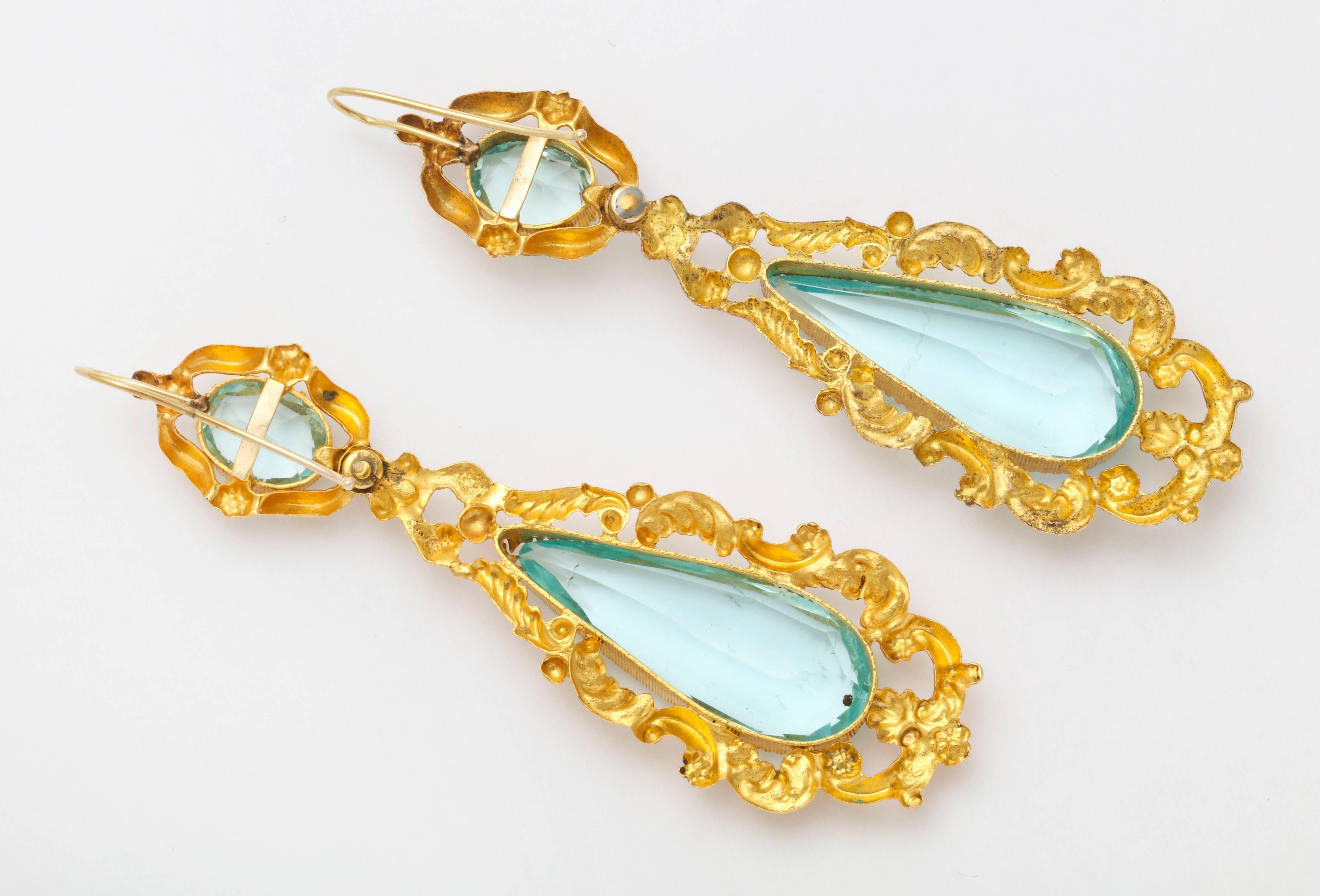 A luscious pair of lightweight chandelier earrings, straight from a 19th century history epic, are ablaze with clear, aquamarine teardrop paste stones surrounded by gold pinchbeck scrolls and vines engraved in repousse, In the early 1800's gold was