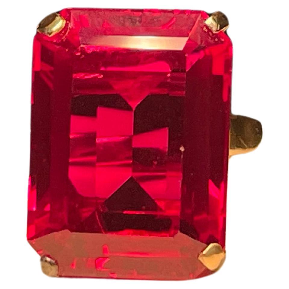 This is a 14K yellow gold Emerald Cut Ruby cocktail ring. The ruby is faceted and set in four gold prongs. Its measurements are 0.75in x 0.5in. The side walls between the prongs are pierced in a rectangular shape. It is signed 14K inside one of the