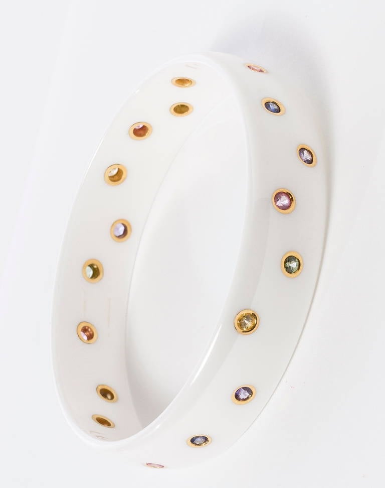 White ceramic dome bangle bezel set with 20 multi-colored sapphires (2.5mm to 3 mm) in 18 karat yellow gold scattered around bracelet.  Sapphire colors in pink, blue, yellow, green & lavender .66ct TW.
Polished finish.