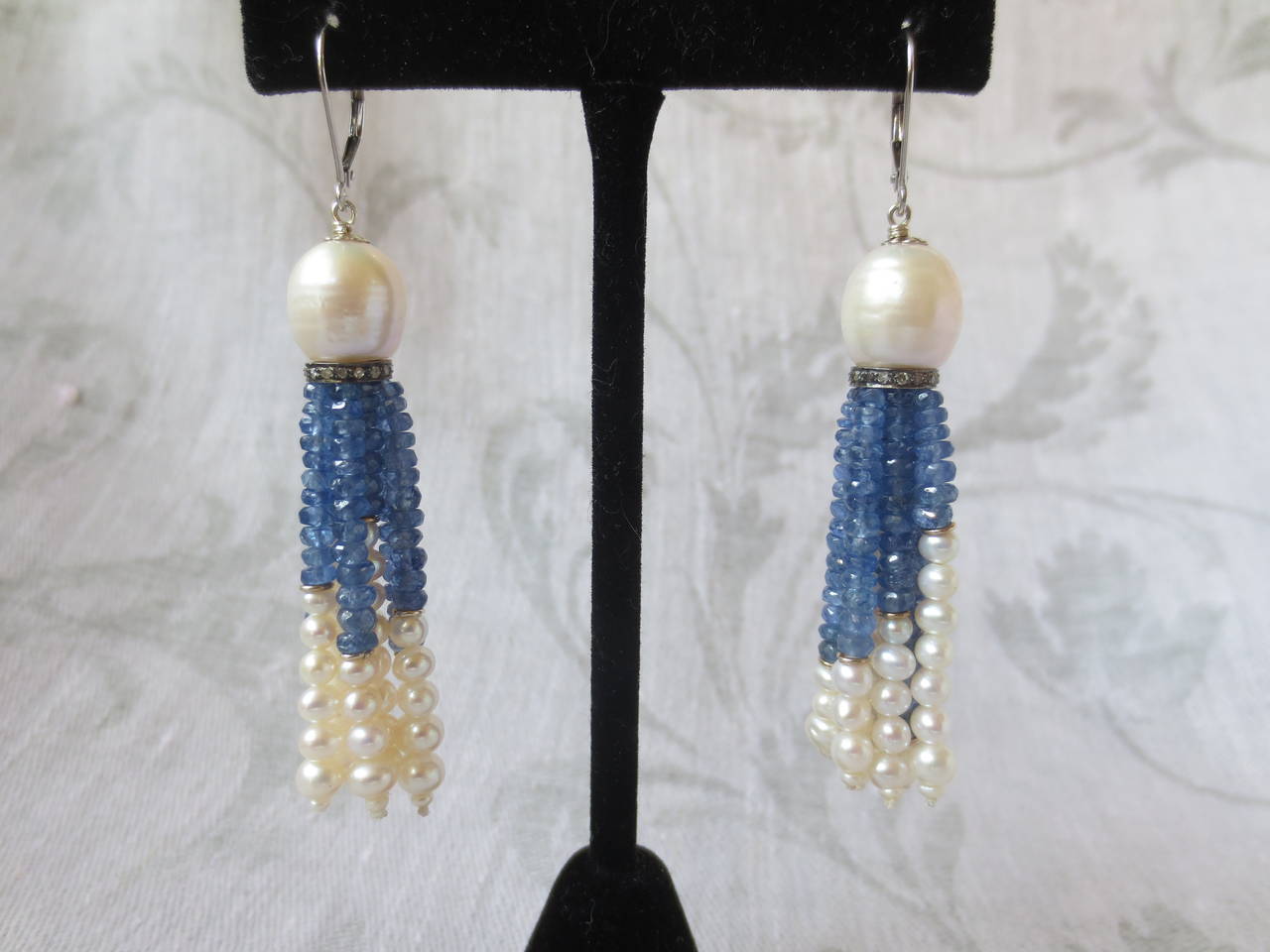 These timeless white pearl and sapphire tassel earrings with 14k white gold lever backs feature a large pearl drop cup set on a sterling silver and diamond encrusted roundel. The tassels are made with faceted blue sapphire beads and graduated