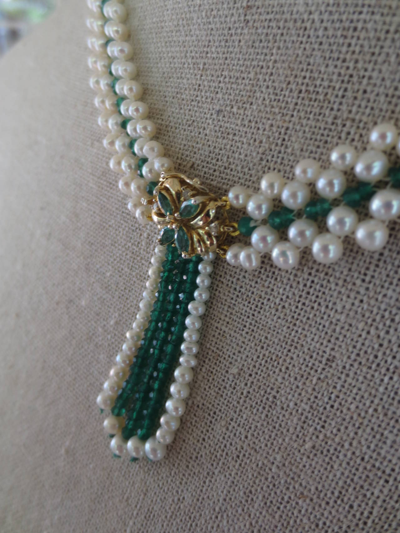 This handwoven pearl, emerald, and 14K yellow gold beaded necklace is accented by a beautiful centerpiece that also serves as the necklace's clasp. The floral centerpiece is made of 14 k yellow gold and accented with emerald stones and small