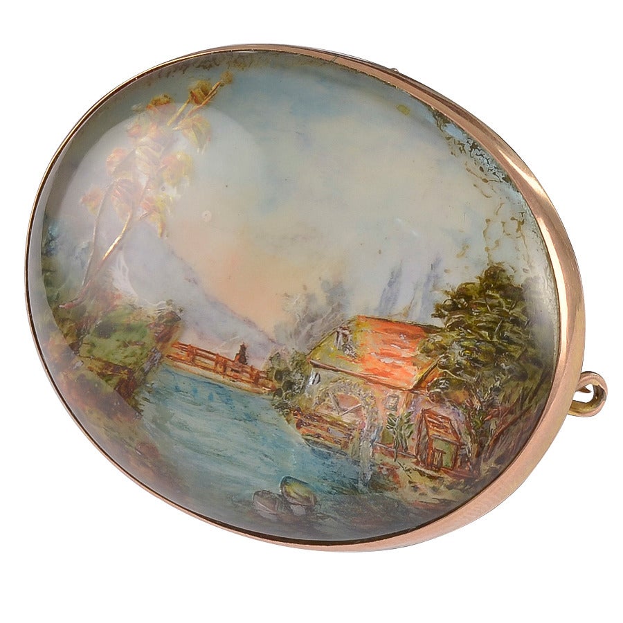 The tranquil oval scene depicting a river, a figure on a bridge,a watermill,distant mountains and perfectly balanced by trees on either bank. 
The frame is unmarked 18k Gold and the Rock Crystal is backed with an oval Silver plaque. A miniature