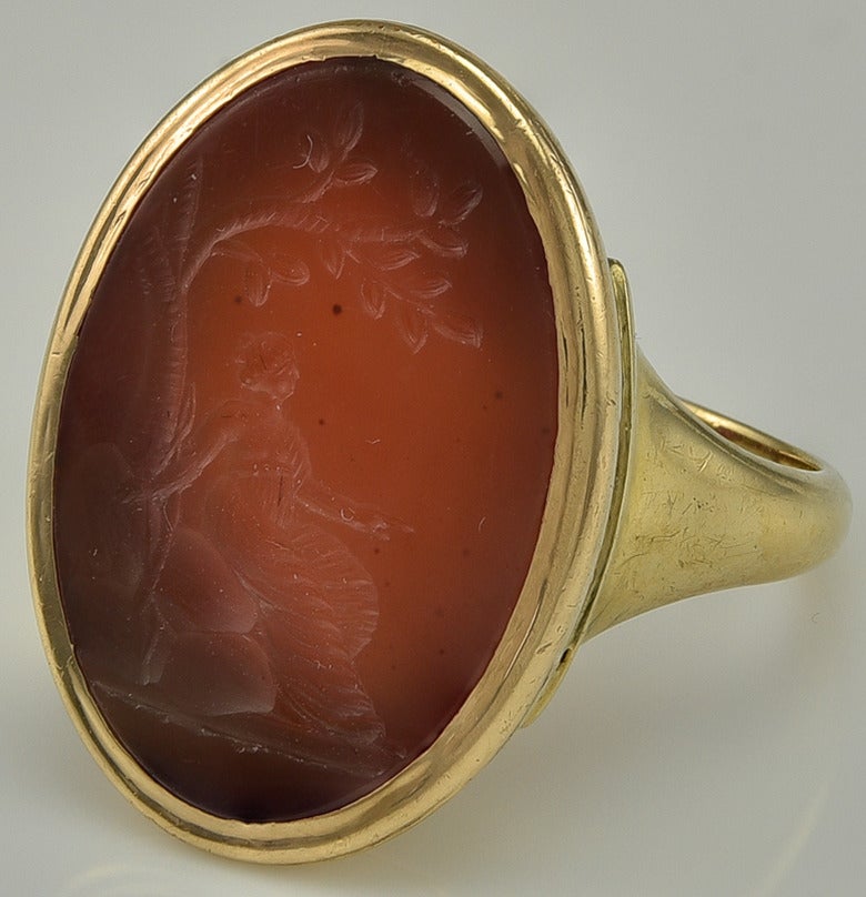 The Carnelian carved with the image of Diana, Goddess of the hunt, seated beneath a tree and pointing, her bow leaning against the trunk. She is clothed in a multi folded Roman stola with her breasts exposed.
The ring mount in unmarked 18k Gold to