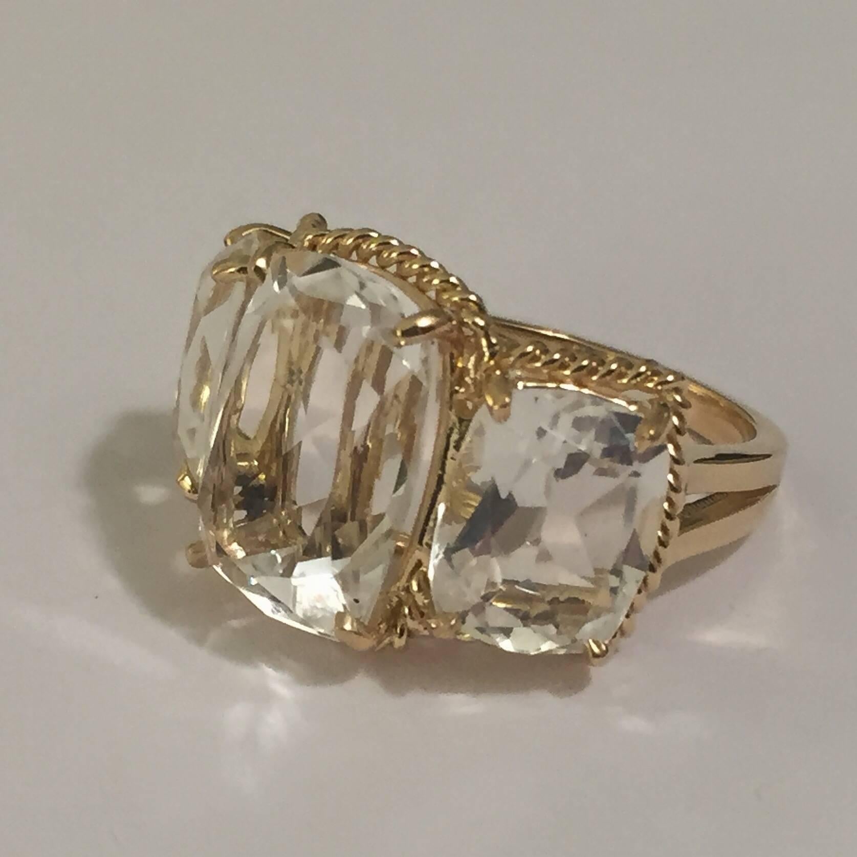 Elegant 18kt Yellow Gold Three Stone Ring with Rope Twist Border with split shank detail. The ring features three faceted cushion cut Rock Crystal stones surrounded by twisted gold rope. The center Rock Crystal stone measures 5/8