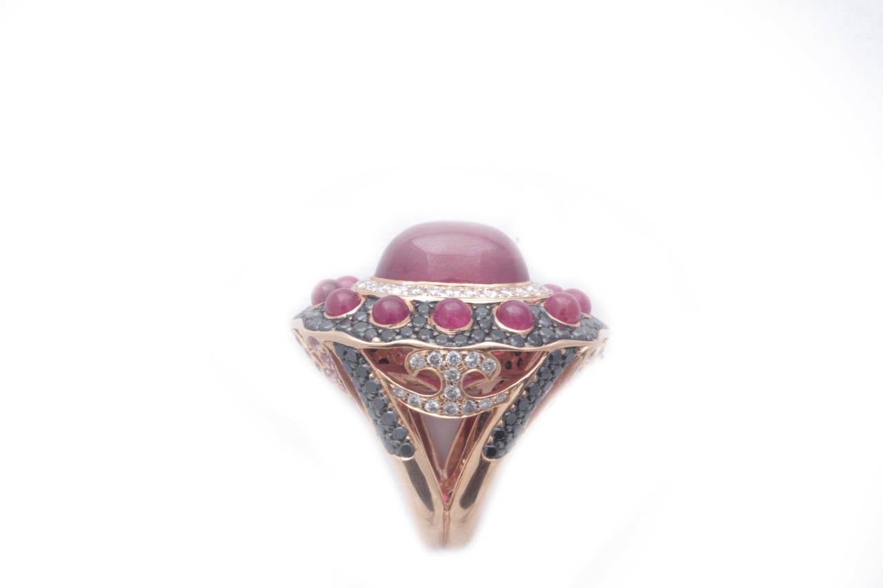 An imposing cocktail ring centering a 12ct cabochon ruby, confined by a circular pavè of brilliant cut diamonds (3ct weight), embellished by an additional waved circumference presenting smaller cabochon rubies on a black diamond pavè. 18kt gold