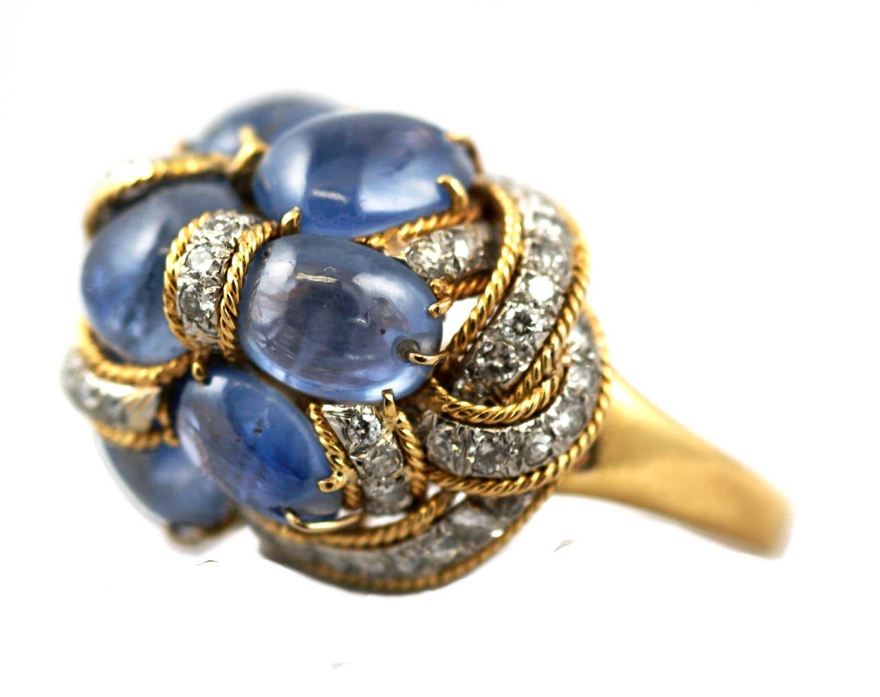 A fun cocktail ring designed as an 18kt yellow gold knot holding together an assemblage of blue cabochon sapphires, embellished by brilliant cut diamonds. Circa 1975.

Adjustable Ring Size