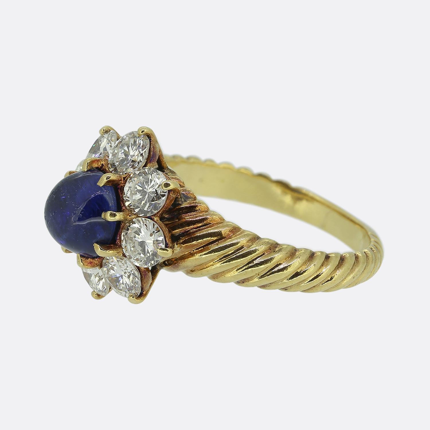 Here we have a striking cluster ring from the renowned French jewellery designer, Mauboussin. At its heart lies a captivating blue cabochon-cut sapphire, encircled by a halo of bright white round brilliant-cut diamonds. These radiant stones are set