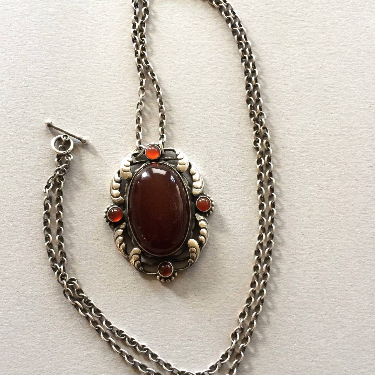 Georg Jensen Pendant No. 158 With Carnelian.

A very rare piece. 

Large deep colored carnelian cabochon center stone flanked with 4 lighter stones. Intricate Foliate design. 29 inch original chain with toggle clasp.

Measures:	2