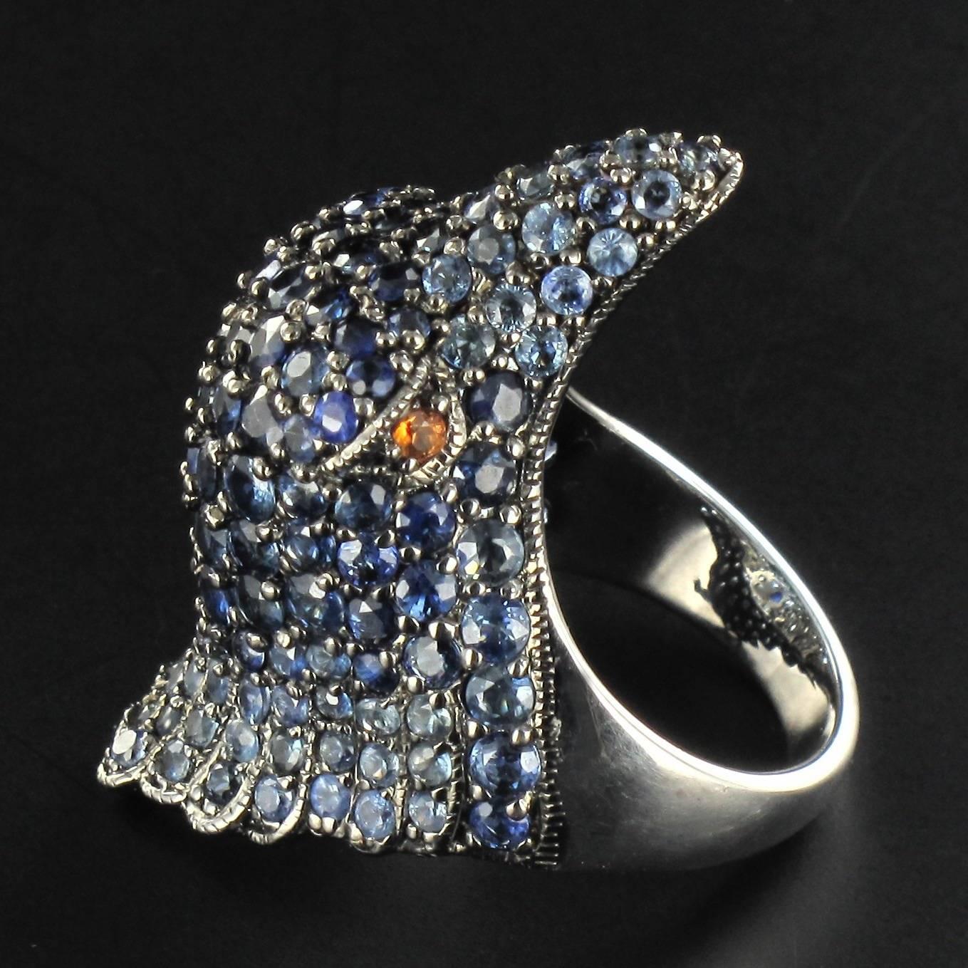 Sterling silver ring.
This surprising ‘animal’ style ring displays the head of an eagle entirely set with numerous sapphires that vary in colour from light blue to dark blue. The piercing eyes are formed of 2 orange sapphires. 
Total weight of