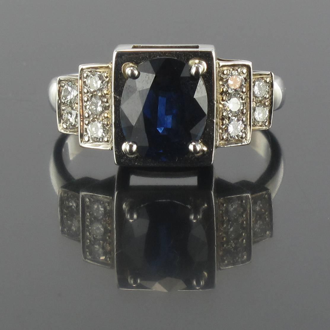 Ring in 18 carat white gold, eagle head hallmark. 

This gorgeous sapphire ring echoes the clear cut geometric lines of the Art Deco style. It is set with a central oval blue sapphire of a deep and uniform blue that is accentuated by 3 plus 2