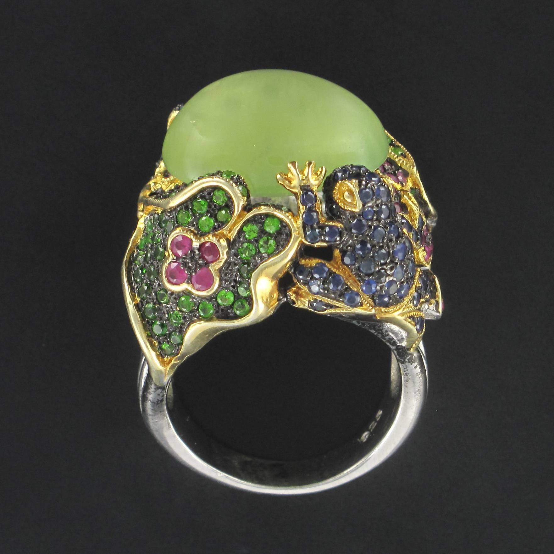 Silver Ring.

This original design ring features a prehnite cabochon held by 2 frogs and 2 water lily pads. The frogs are set with a multitude of tiny round sapphires and the water lily leaves with tsavorite garnets, the lily flowers are set with
