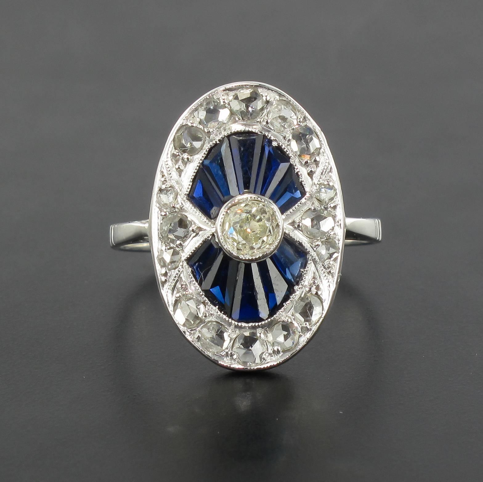 Ring in 18 carat white gold, eagle head hallmark.

This radiant antique oval shaped ring is bezel set with a central antique brilliant cut diamond surrounded by calibrated sapphires and an outer circle of good sized rose cut diamonds. 
The ring