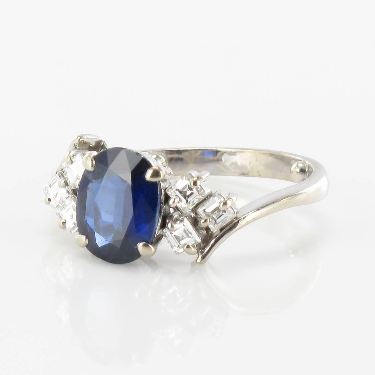 Ring in 18 carat white gold, eagle head hallmark.

This gorgeous white gold ring is claw set with an oval sapphire featuring 3 princess cut claw set diamonds at each side. 
The beginning of the ring band is asymmetric.  

Total weight of the