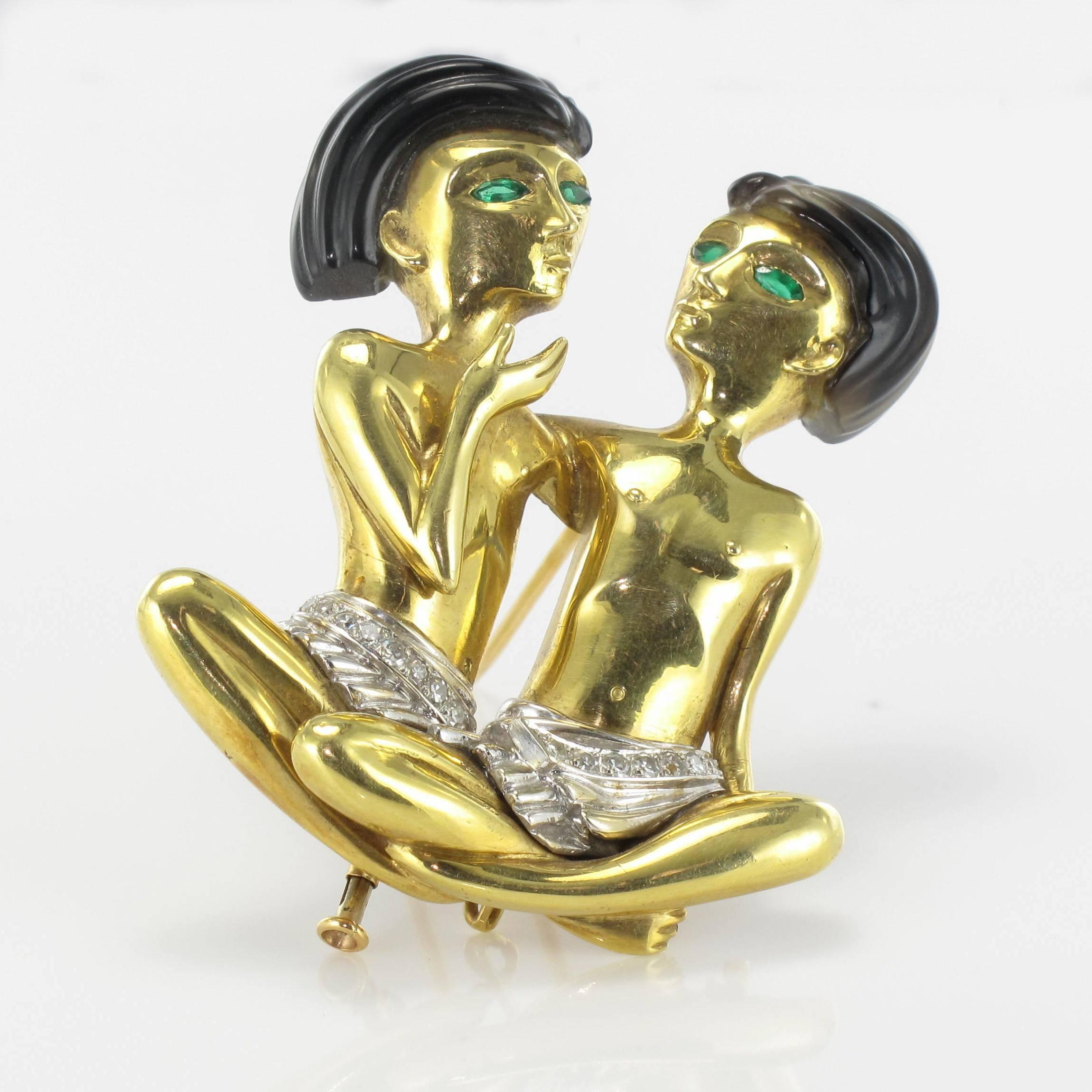 Brooch - Pendant in 18 carat yellow and white gold. 

Featuring 2 small seated Egyptian style figures with emerald eyes. Their hair is agate and their white loincloths are set with rows of diamonds. The clasp has 2 pins with a tube lock. This