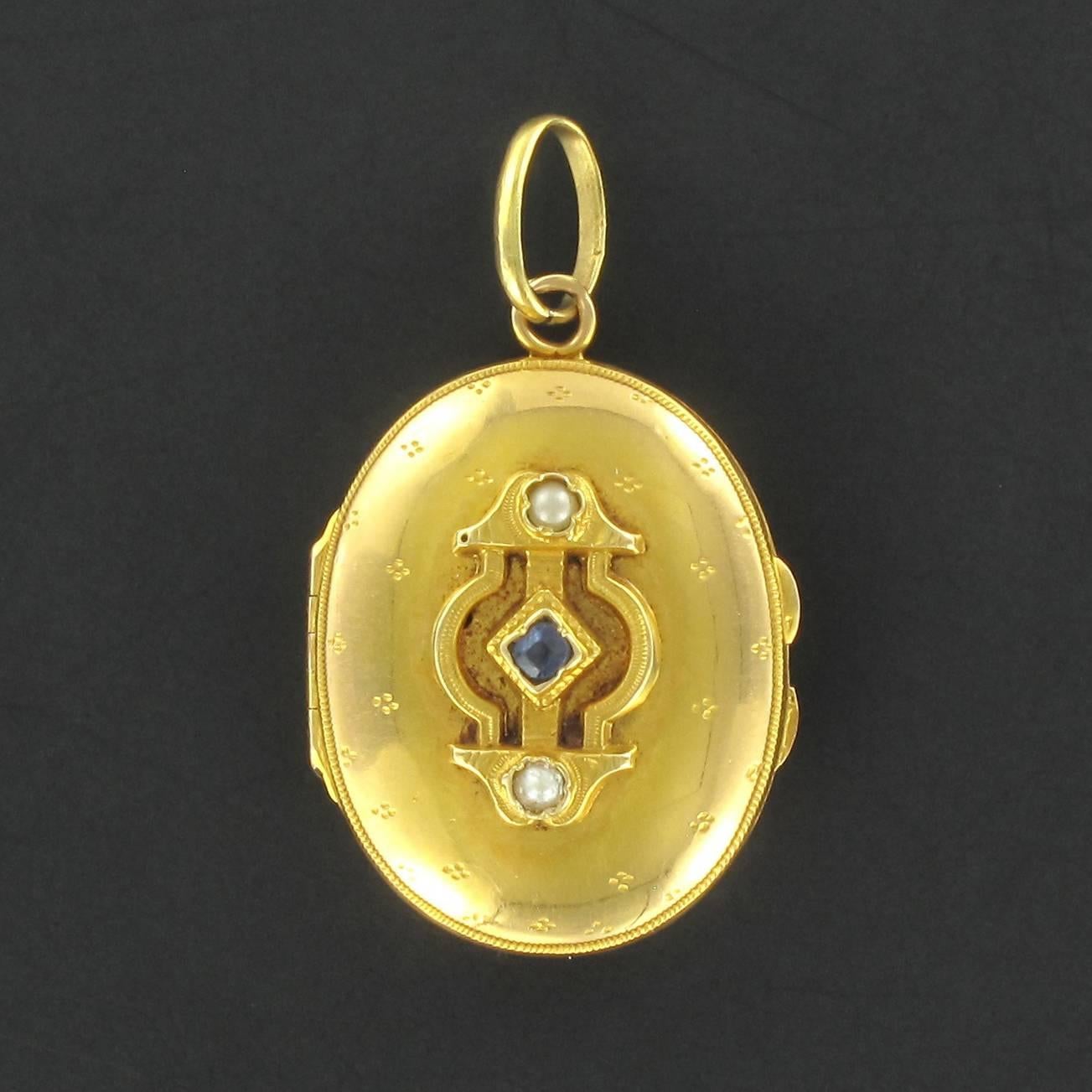 Pendant in 18 carat yellow gold. 

An absolutely charming piece of jewellery, this oval gold locket displays a motif in a chiselled cartouche with a central bezel set blue sapphire and a fine half pearl above and below it. The front and back of the