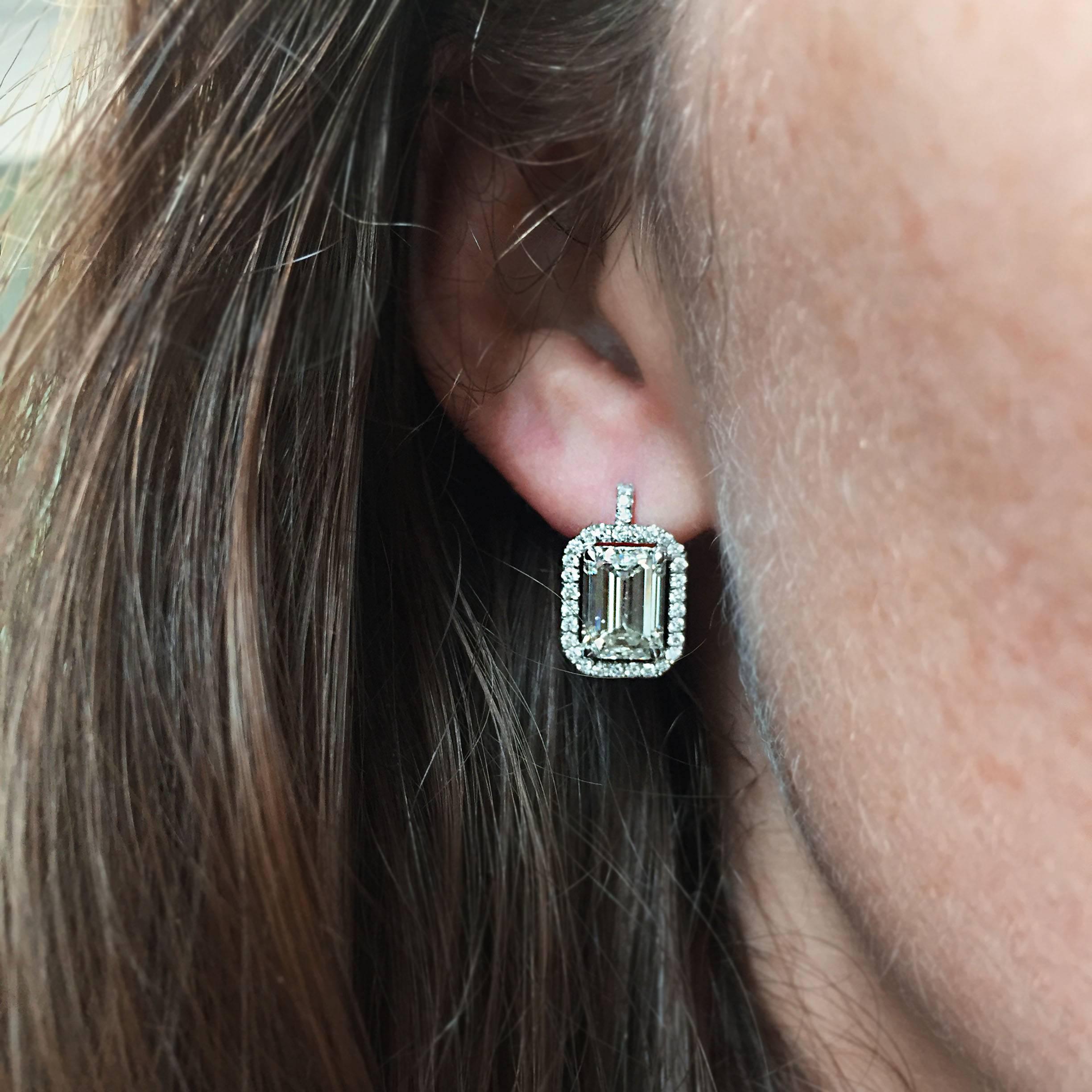 A lovely pair of 18k white gold drop earrings feature two emerald-cut diamonds prong set in a pave halo design. The two large diamonds have I color and VS2 clarity, and weigh 3.24 carats and 3.58 carats, respectively. Earrings contain 62 round-cut