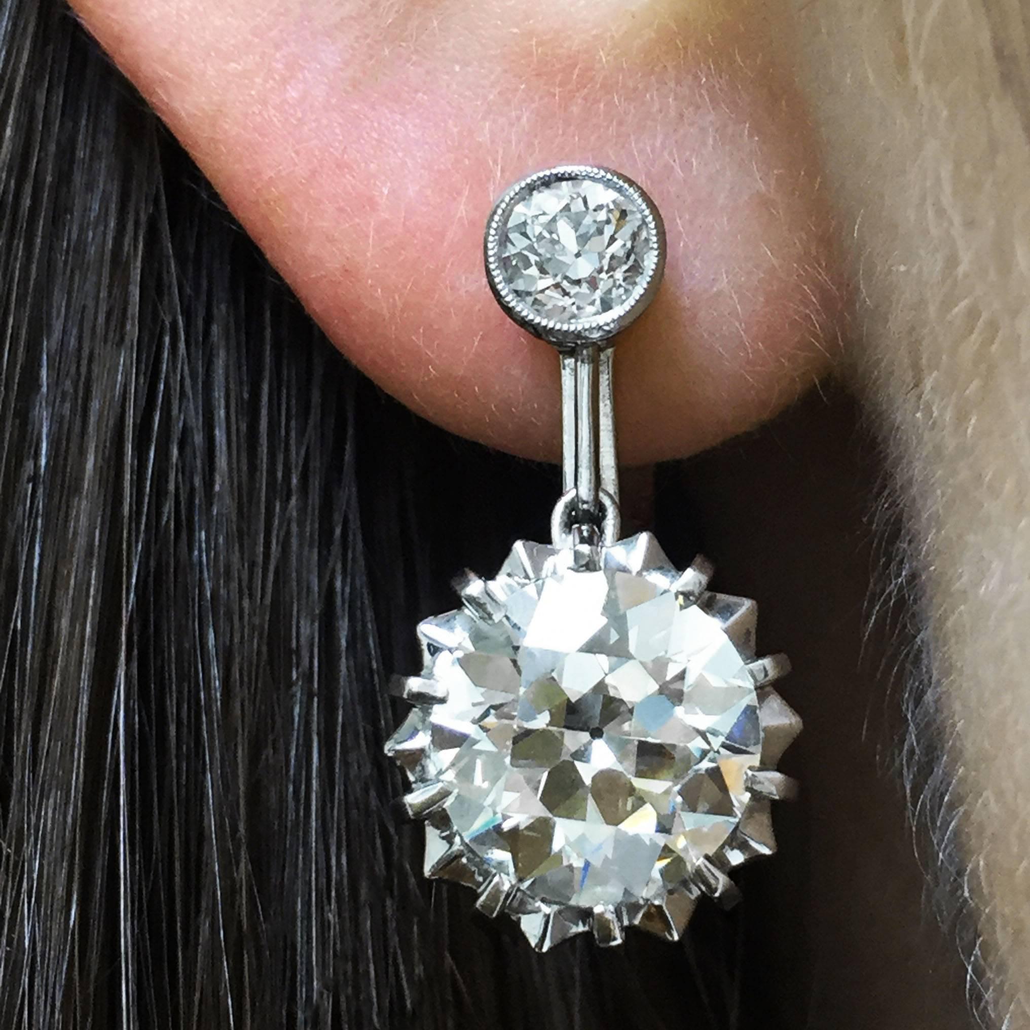 A pair of lovely vintage 18k white gold earrings feature two large Old European-cut diamonds set in unique star-like mountings suspended from smaller bezel-set Old European-cut diamond studs. The larger diamonds weigh 2.81 carats and 2.80 carats and