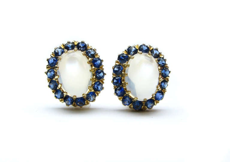 These lovely earrings feature oval cabochon moonstones set in 14kt yellow gold, and are surrounded by a frame of round brilliant blue sapphires. These unique gemstone earrings are a great statement piece for any occasion.