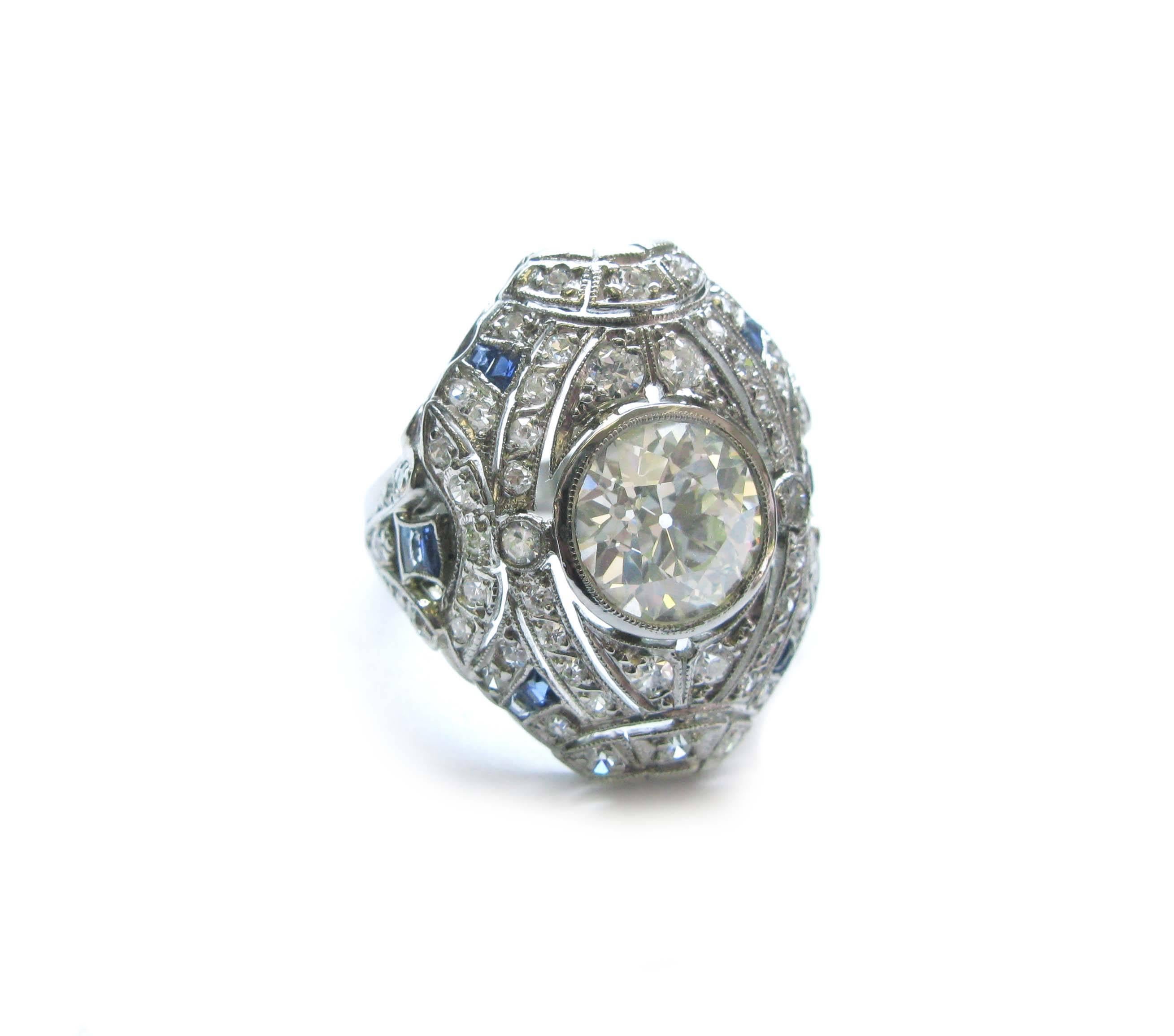 This truly unique 18kt white gold Art Deco ring features a bezel set 2.26ct, K color, VS2 clarity, Old European cut diamond center stone surrounded by two bezel set diamonds and rows of pave set diamonds with blue sapphire accents. This intricately