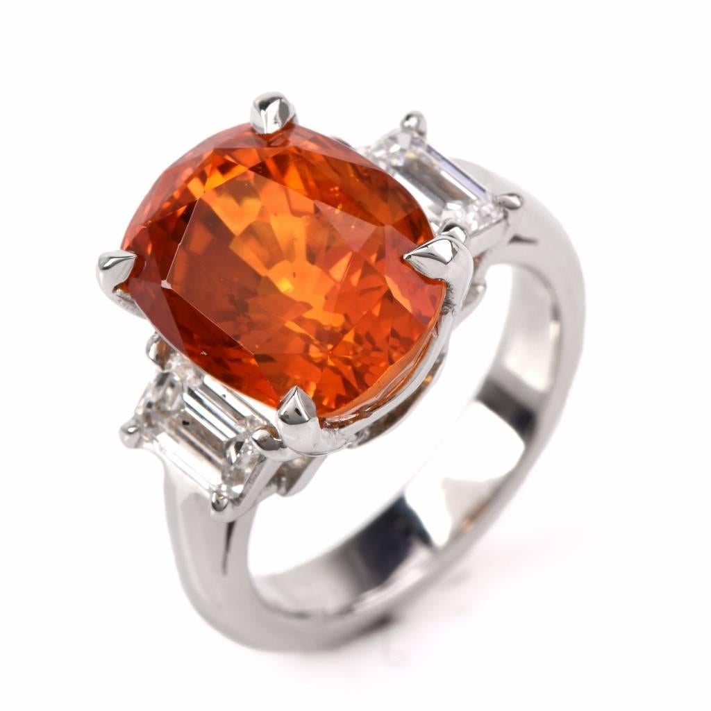 This estate 3-stone ring with a genuine orange sapphire and a pair of emerald-cut diamonds is craftd in solid platinum.The genuine cushion-cut  sapphire weighing 15.04 cts and measuring 12.54 x 10.02 x 9.50 mm is secured by four prongs, atop an