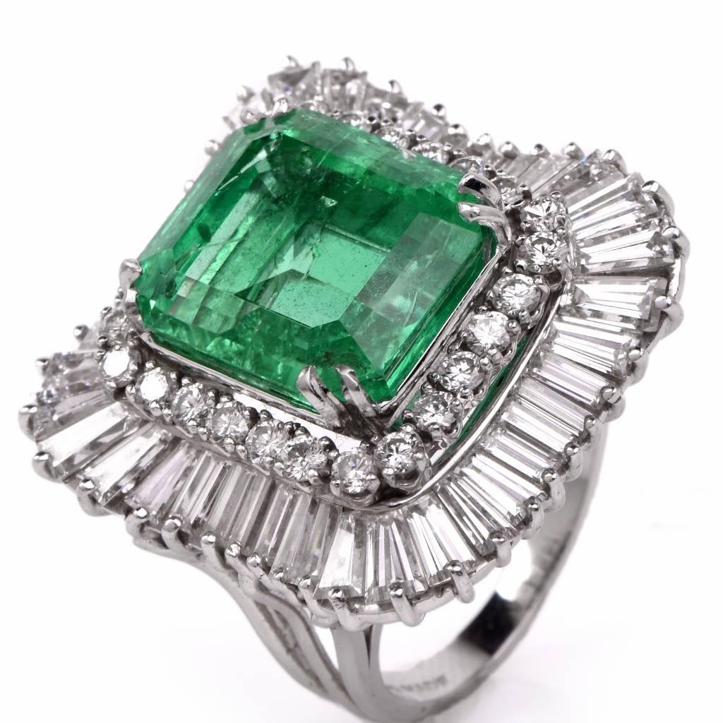 This opulent cocktail ring with an eminent and genuine emerald-cut emerald  is crafted in solid platinum and weighs 26.9  grams. The centrally positioned vibrant transparent emerald weighs approx. 12.36 cts and measures 15.3 mm x 13.3 mm  secured by