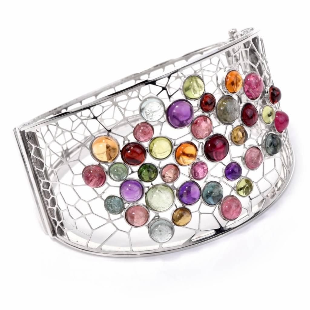 This lovely Italian bangle bracelet displays an open work geometrical design artfully embellished by a cluster of 37 round cabochon multi gemstones (Aquamarine, Garnet, Amethyst, Tourmaline, Citrine, & Peridot) weighing approx. 21.00