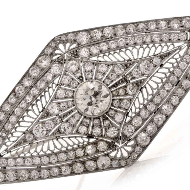 This filigree brooch is crafted in platinum, weighs approximately 19.5 grams.  Designed as a diamond-encrusted lozenge plaque, this Art Deco brooch depicts artistically handcrafted filigree profiles of asymmetric format. Centered with an