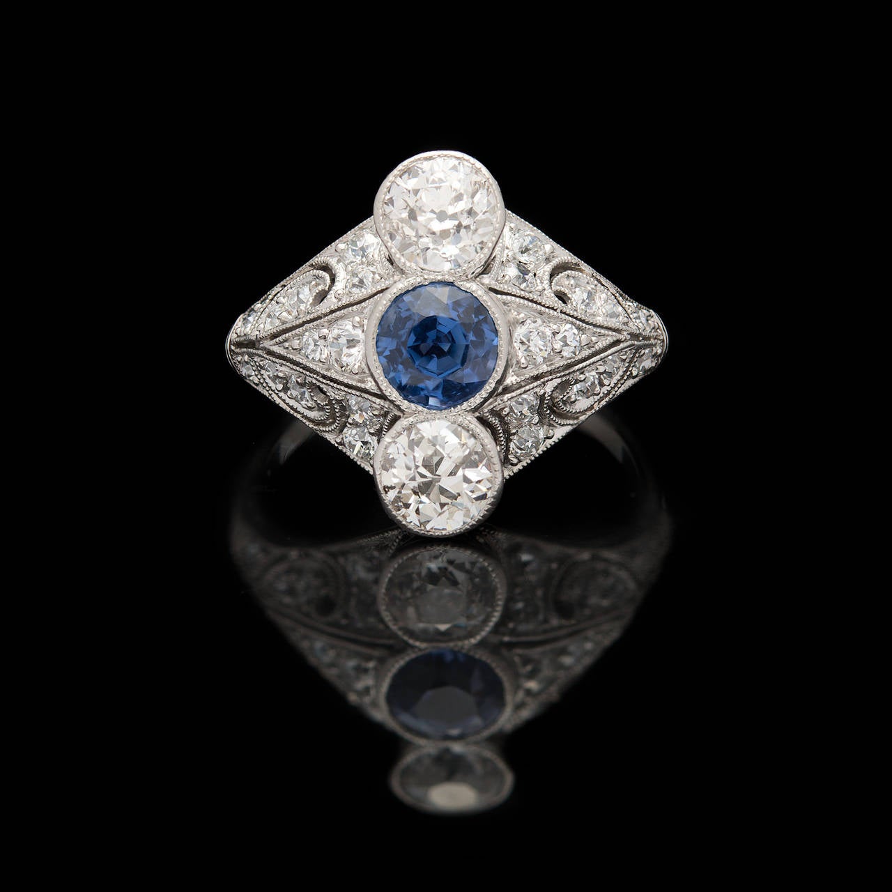 Edwardian Platinum Setting Features a 0.70ct Round Cut Blue Sapphire Flanked by 2 Old European Cut Diamonds and Enhanced by an additional 24 Round Cut Diamonds. The total approximate diamond carat weight is 1.30cts. The ring is a size 6.0 and weighs