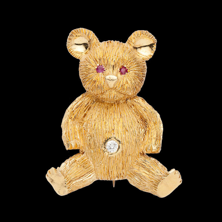 Charming Teddy Bear Brooch in 18Kt Yellow Gold Features 2 Faceted Dark Pink Sapphire Eyes and a Round Brilliant Cut Diamond in the center of its belly. The Diamond is approximately 0.06ct. The brooch measures 1.4 x 1.1 inches and weighs 12.2 grams.