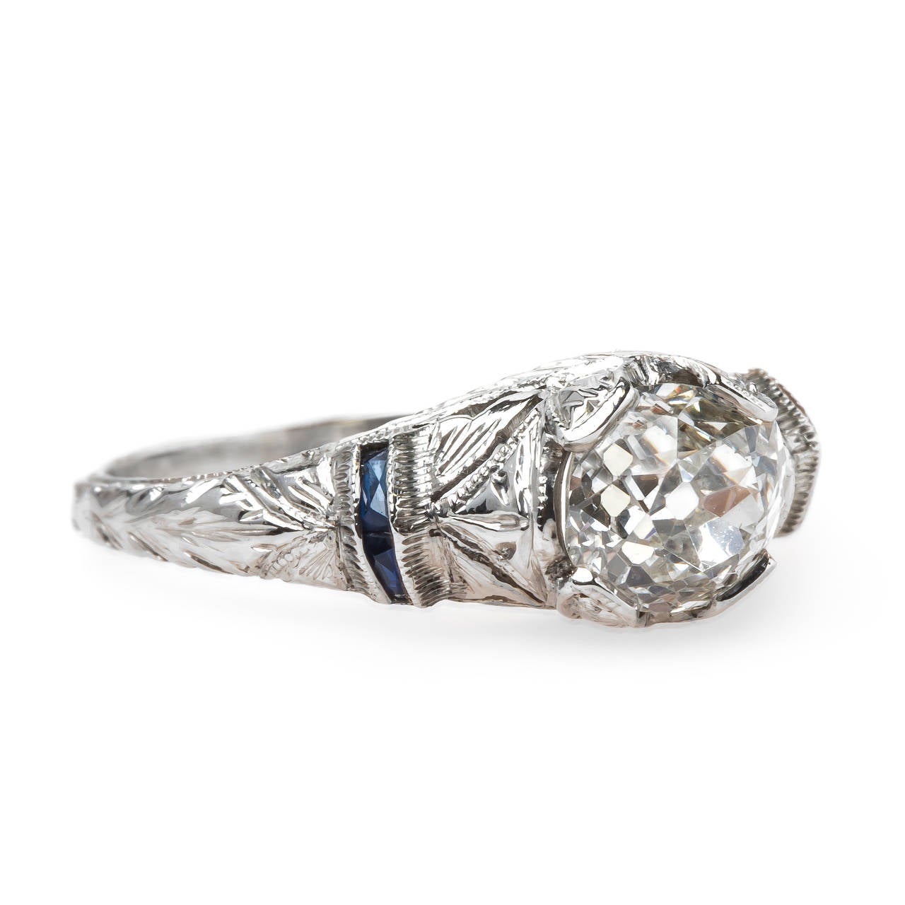 Edenstone is a delicate and authentic Edwardian era (circa 1915) platinum engagement ring featuring a 0.99ct EGL certified Old Mine Cushion Cut diamond graded I color and VS2 clarity. The center stone is nestled between four French Cut bright blue
