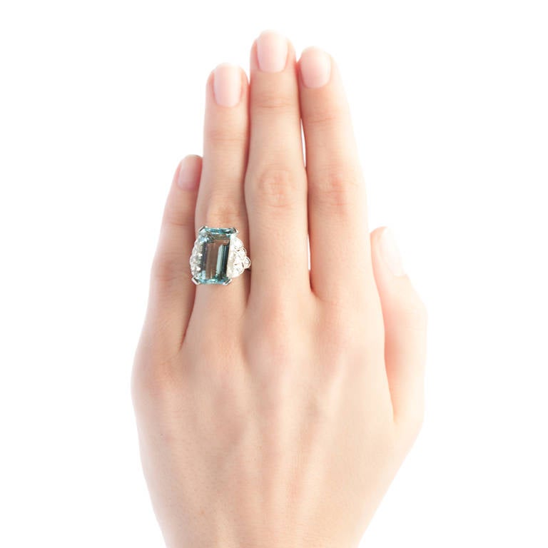 Valley Falls is a show-stopping late Art Deco era platinum set cocktail ring centering a stunning 10.26ct Retangular Step Cut natural aquamarine. Flanked on either side by decadent, scalloped stepped-down shoulders and studded with twelve Single Cut