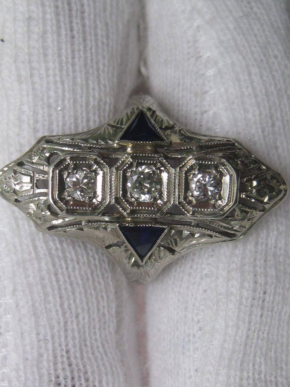 This wonderful fashion ring from 1920s Art Deco features three diamonds flanked by synthetic sapphires set in 18K white gold filigree. The diamonds hold a 0.20 cttw. with an H color and a VS1 clarity. Transport yourself to a time of high fashion