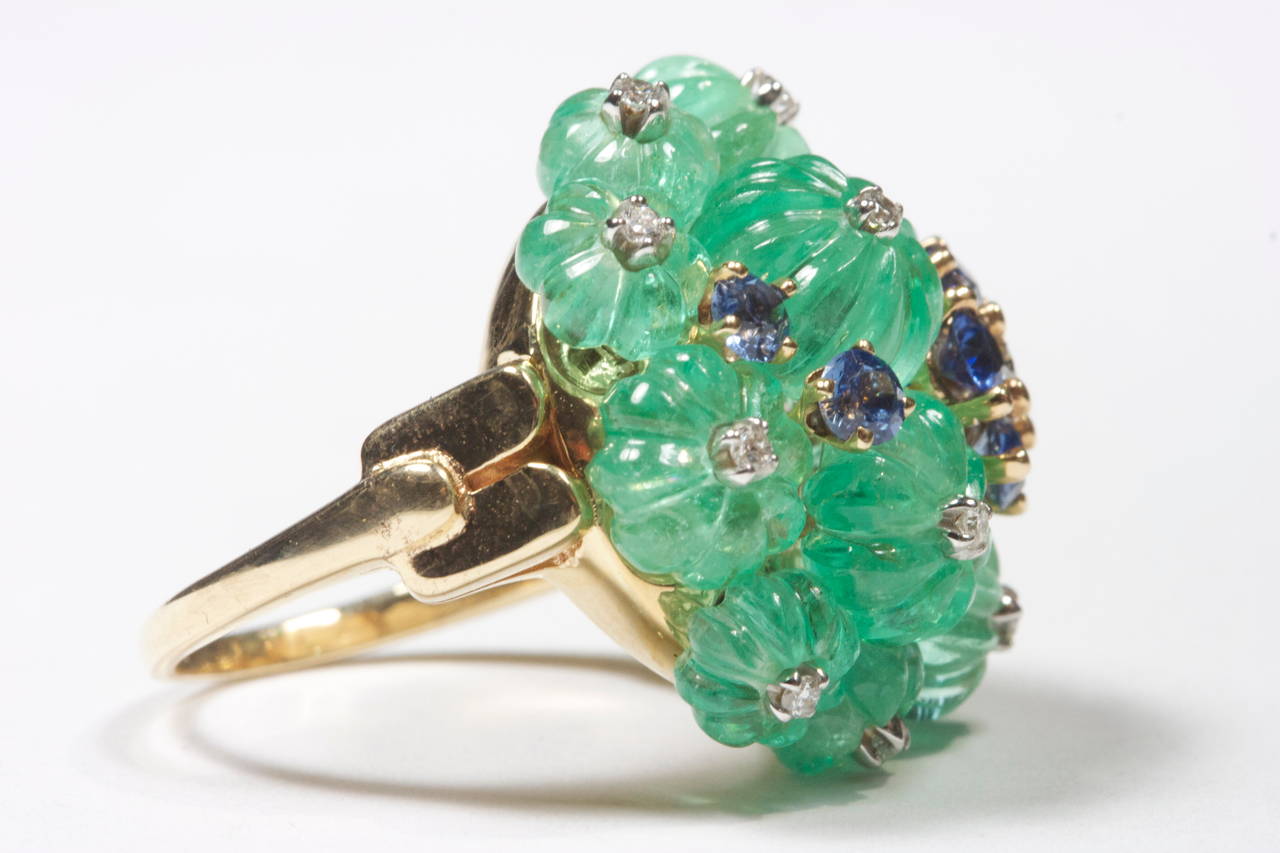 A beautifully designed ring portraying a bouquet of flowers. The ring is created with carved emerald, round blue sapphires and clean white diamonds. Crafted in 18k gold.

Ring Size 5 3/4