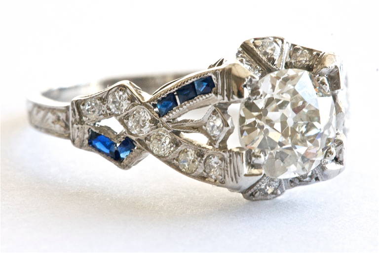 Charming art deco ring with a 1.15 carat diamond and accented by sapphires and diamonds. Graceful and romantic. We grade the diamond as H color and VS clarity.