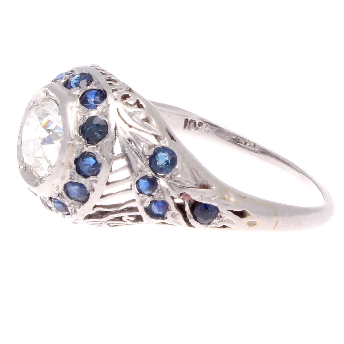 An imaginative art deco design that features iconic style from this era. Designed with a lively center diamond which is surrounded by a halo of deep blue sapphires. Accented with stylish geometric carved filagree lines and more well matching deep