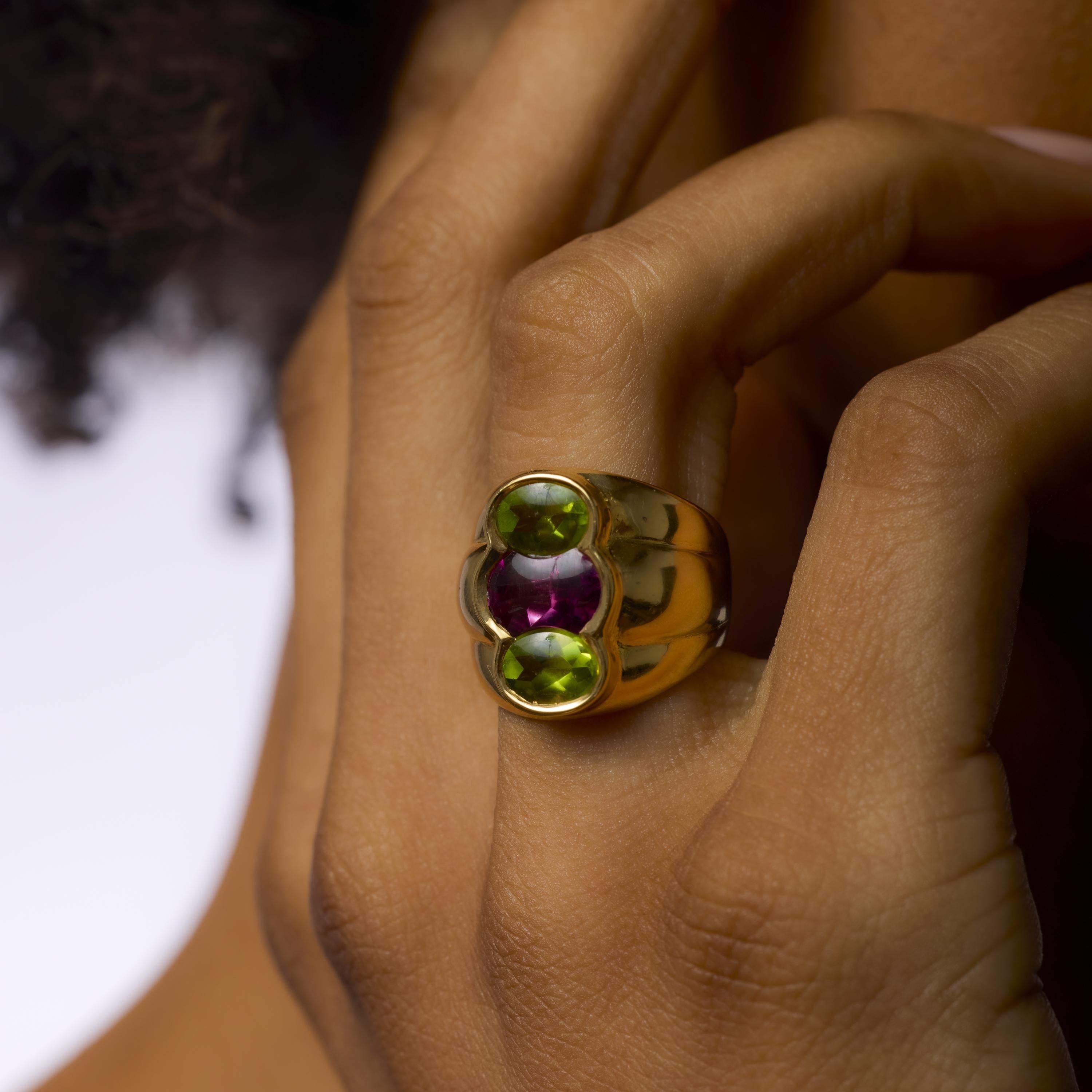 Questo vibrante anello vintage degli anni '80 mette in mostra due peridoti verdi dal taglio ovale che affiancano una ricca ametista viola al centro. Incastonato in un luccicante oro giallo 18 carati, l'audace contrasto di colori irradia una giocosa
