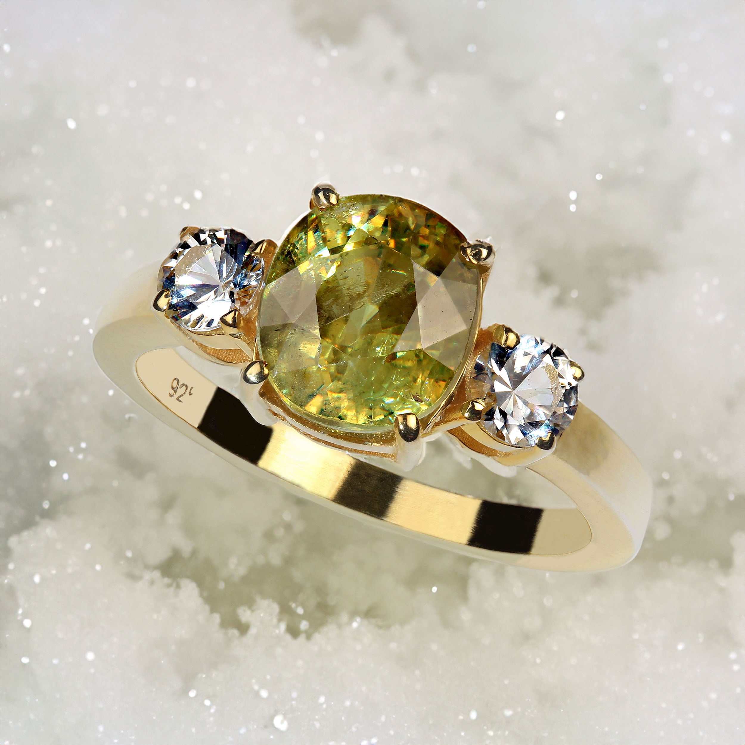 Size 8 ring of oval Sphene and white Sapphires.  This classic ring features a scintillating limey yellowy oval Sphene of 3.35ct accented with two round 4MM white Sapphires of 0.67ct. These lovely gemstones are custom set in gold rhodium over