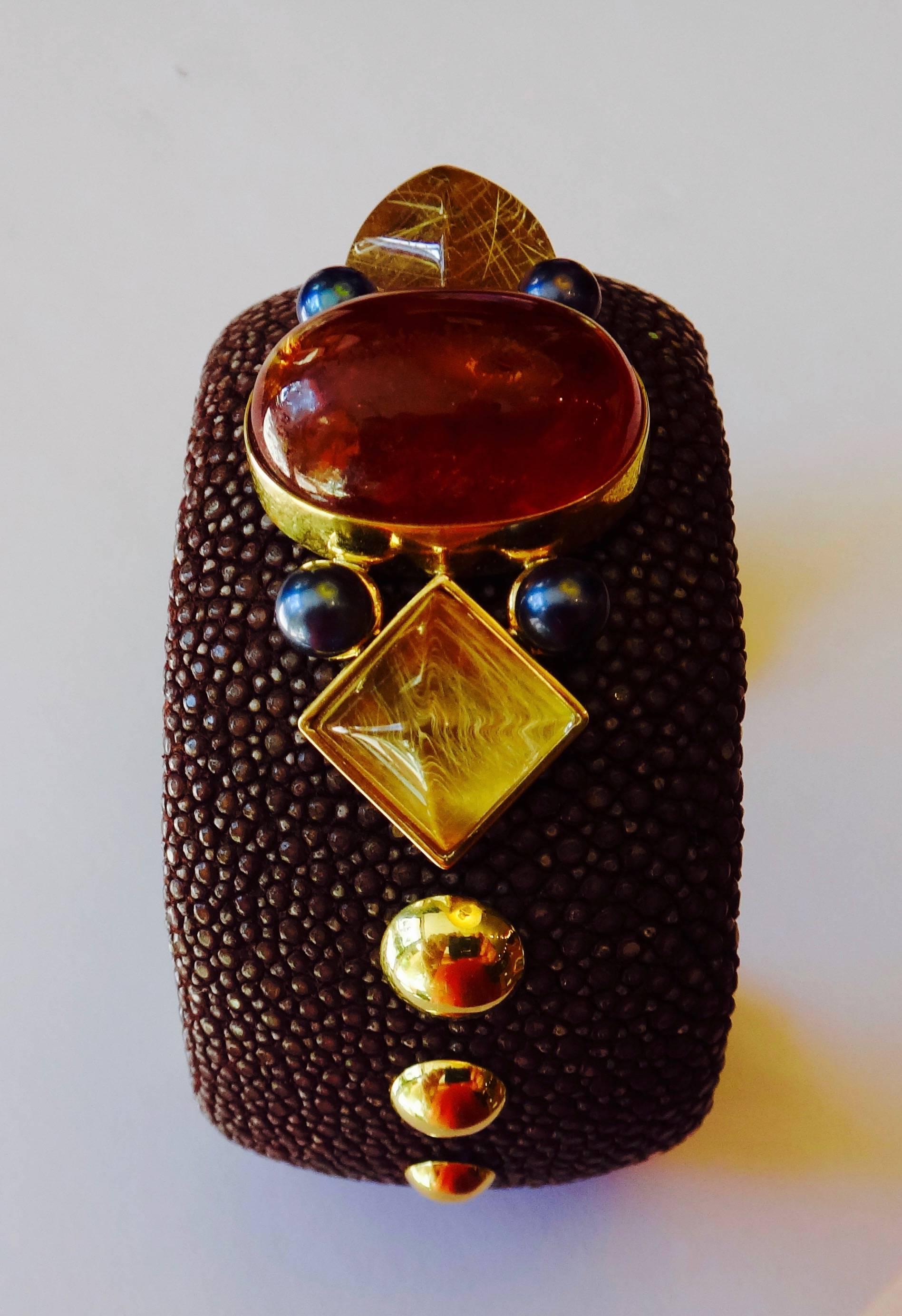 Mocha colored stringray cuff is decorated with a large cabochon hessonite garnet, 2 sugarloaf cabochons of rutilated quartz and black Akoya pearls.  All set in 18k yellow gold.  Inside diameter is 6.75 inches.  There is a sprung steel core within
