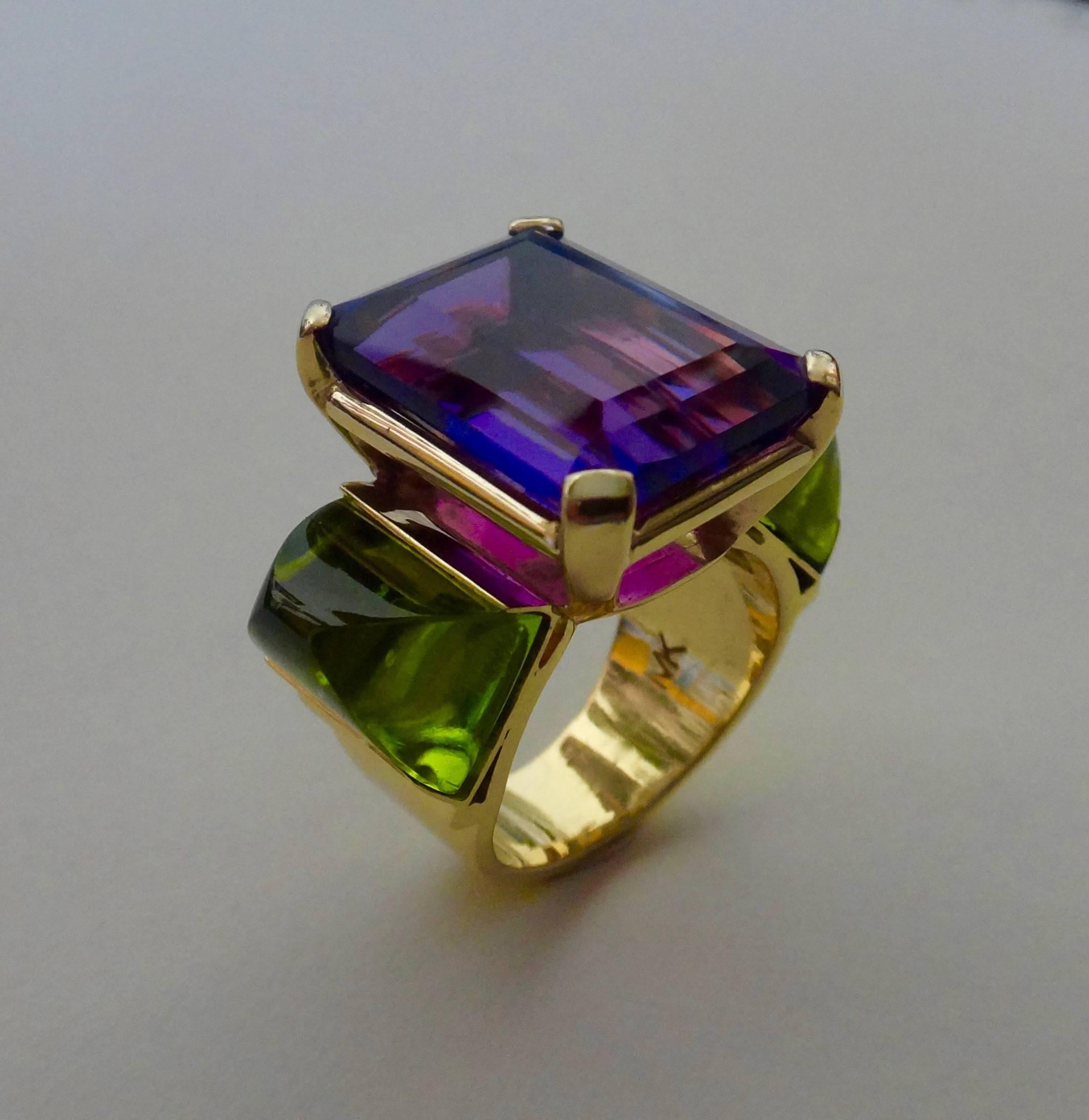 Set in this hand fabricated 18k yellow gold ring is an emerald cut African amethyst flanked by a pair of perfectly matched, sugarloaf cabochon peridots cut in Idar Oberstein, Germany.  This ring is perfect for someone with a knuckle problem as the