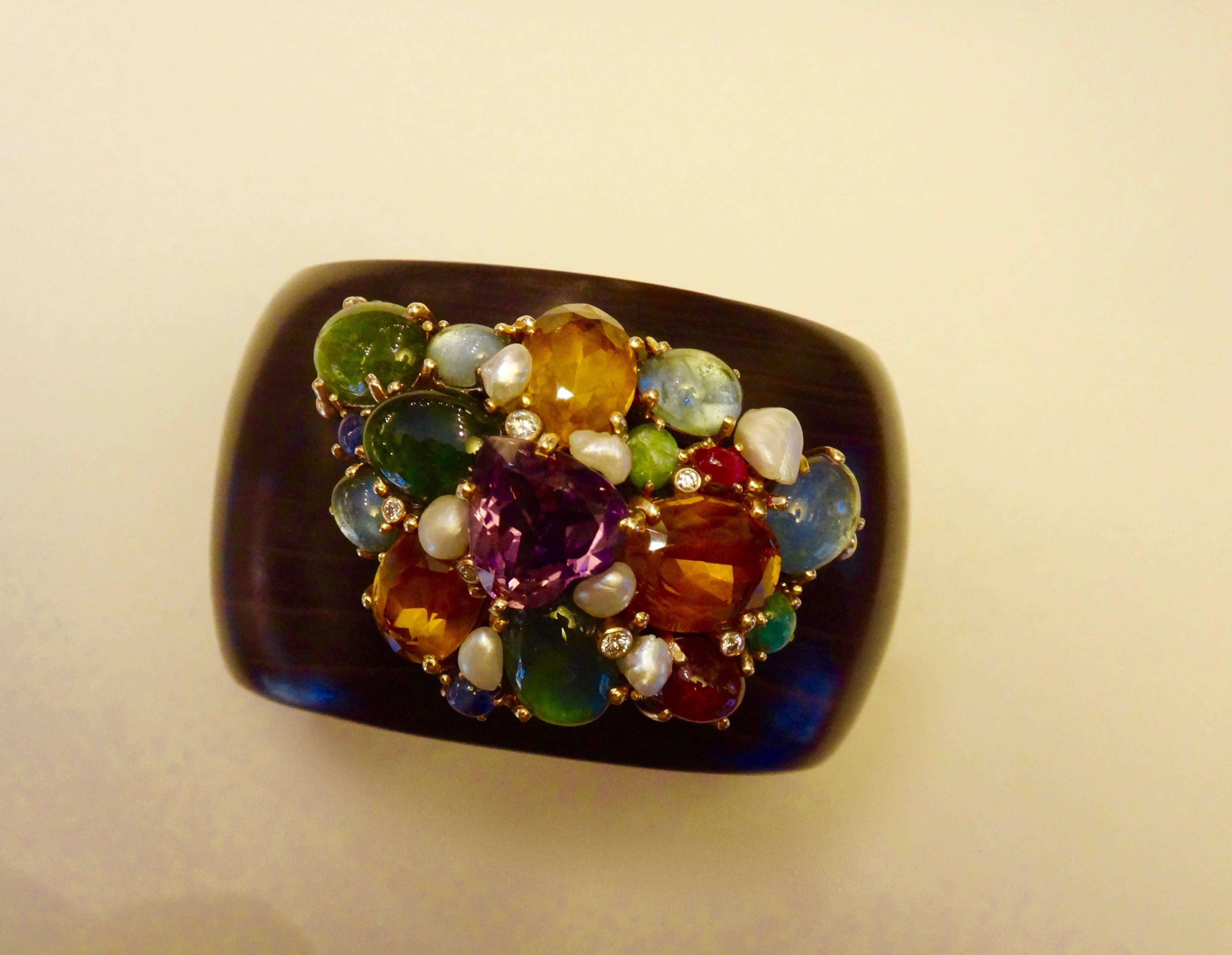 A collection of faceted and cabochon gems including amethyst, citrine, green tourmaline, blue sapphire, and emerald are clustered together on a carved ebony cuff bracelet.  Also included in this bold and dramatic cuff are natural freshwater pearls