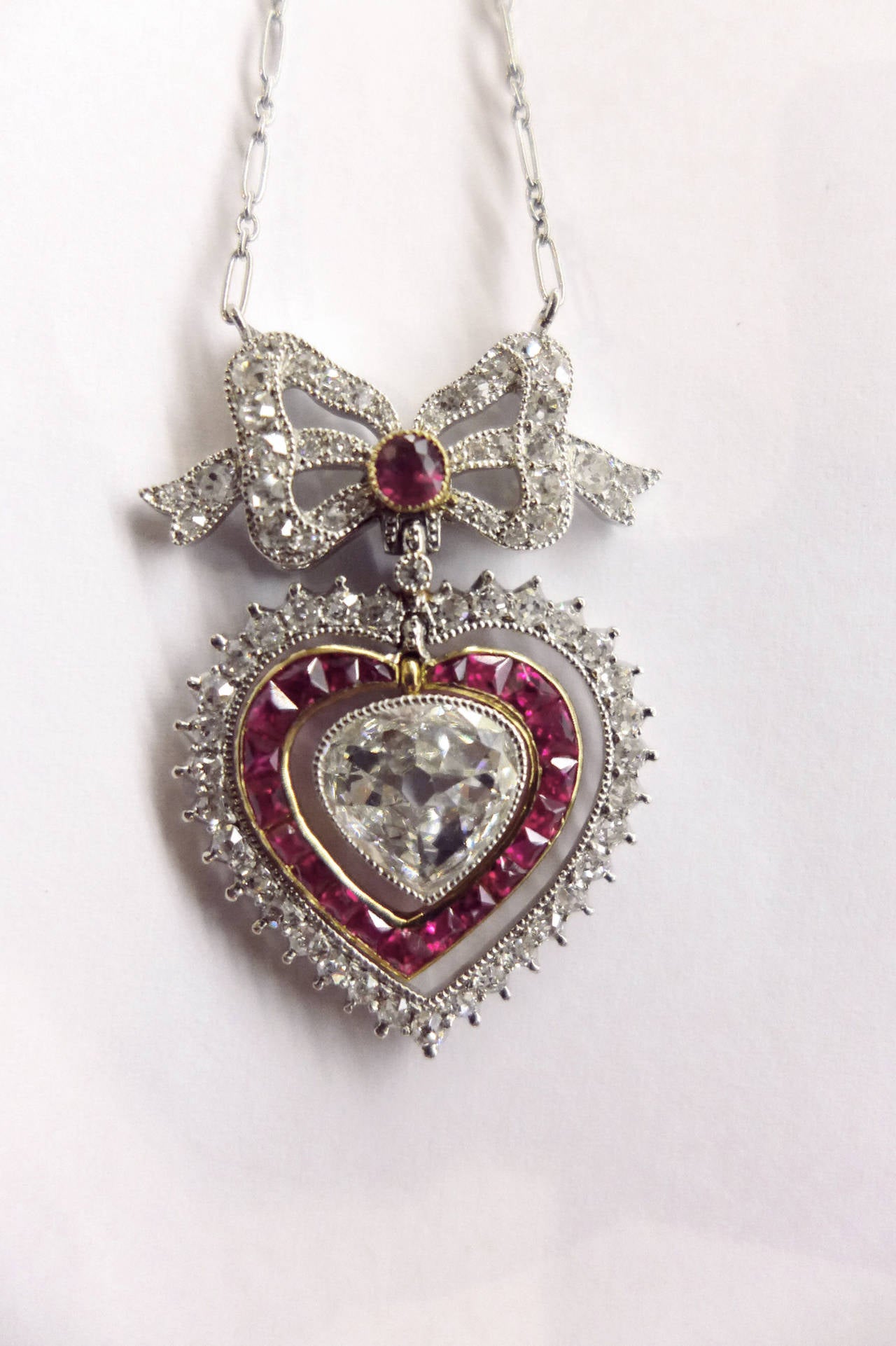 A platinum charming necklace set with an old cut diamond heart-shaped surrounded by diamonds and calibré cut rubies set in yellow gold. 
Above the heart, a bow set with a ruby and diamonds. 
Pendant width : 40 mm 
Heart diamond weight : about 1