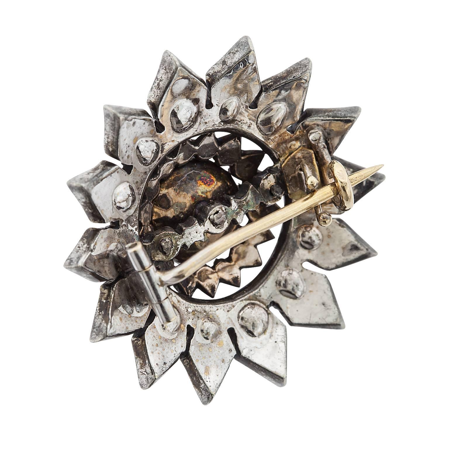 A Victorian era silver over gold cabochon moonstone and rose cut diamond brooch.  The moonstone is partially bezel set, sitting atop a single row of surrounding diamond, framed by a floral pattern of two rows of diamonds.  The moonstone measures