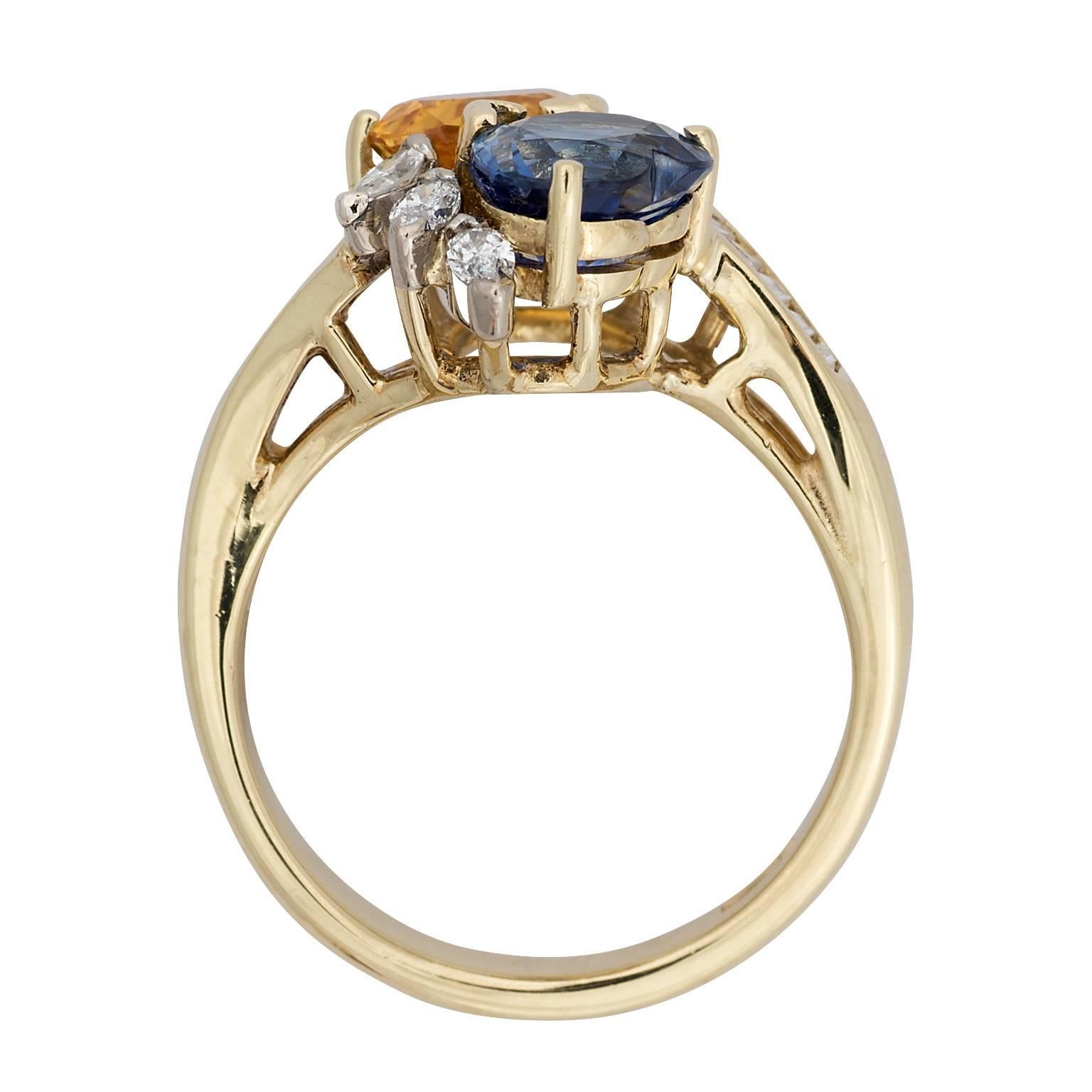 A Hammerman heart shaped orange and blue sapphire and diamond 18k yellow gold ring.  The two sapphires have a total weight of 2.00 carats.  The ring is designed as a bypass with three marquise diamonds on the side of each sapphire, and a channel set