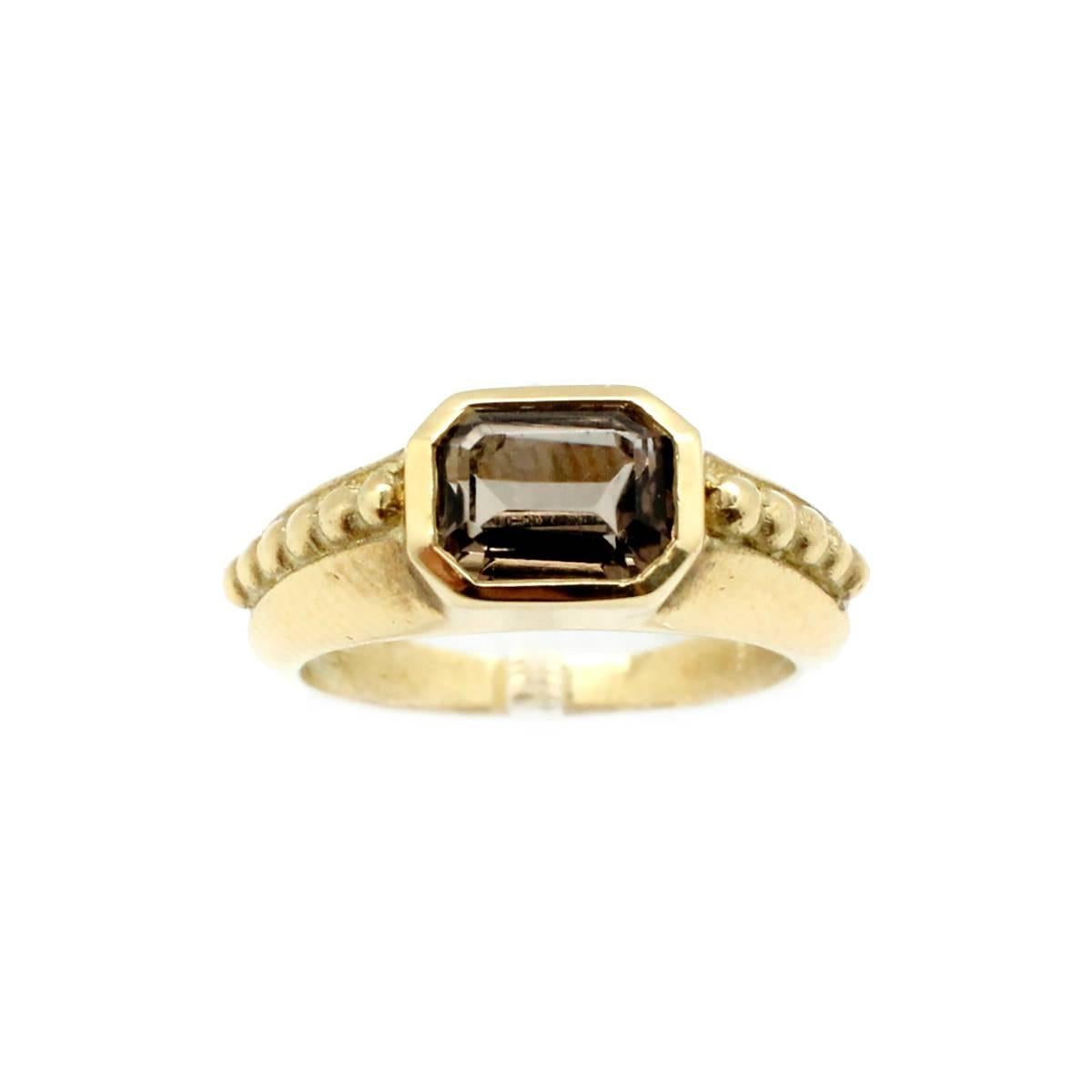 This ring is made in 18k yellow gold by Judith Ripka. The piece is signed “© Judith Ripka 18k.” The ring holds a single piece of smoky quartz at its center. The ring measures 7mm wide, and it weighs 7.5 grams. It is a size 6.