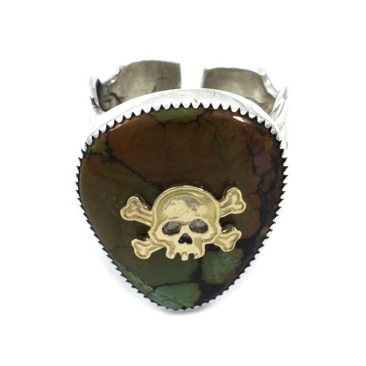 This ring is made in sterling silver by Kit Carson. The ring features a gold skull set on top of an orange-green turquoise stone shaped like a guitar pick. The ring measures 22x26mm, and it weighs 21.0 grams. The ring is size 9. It is signed 