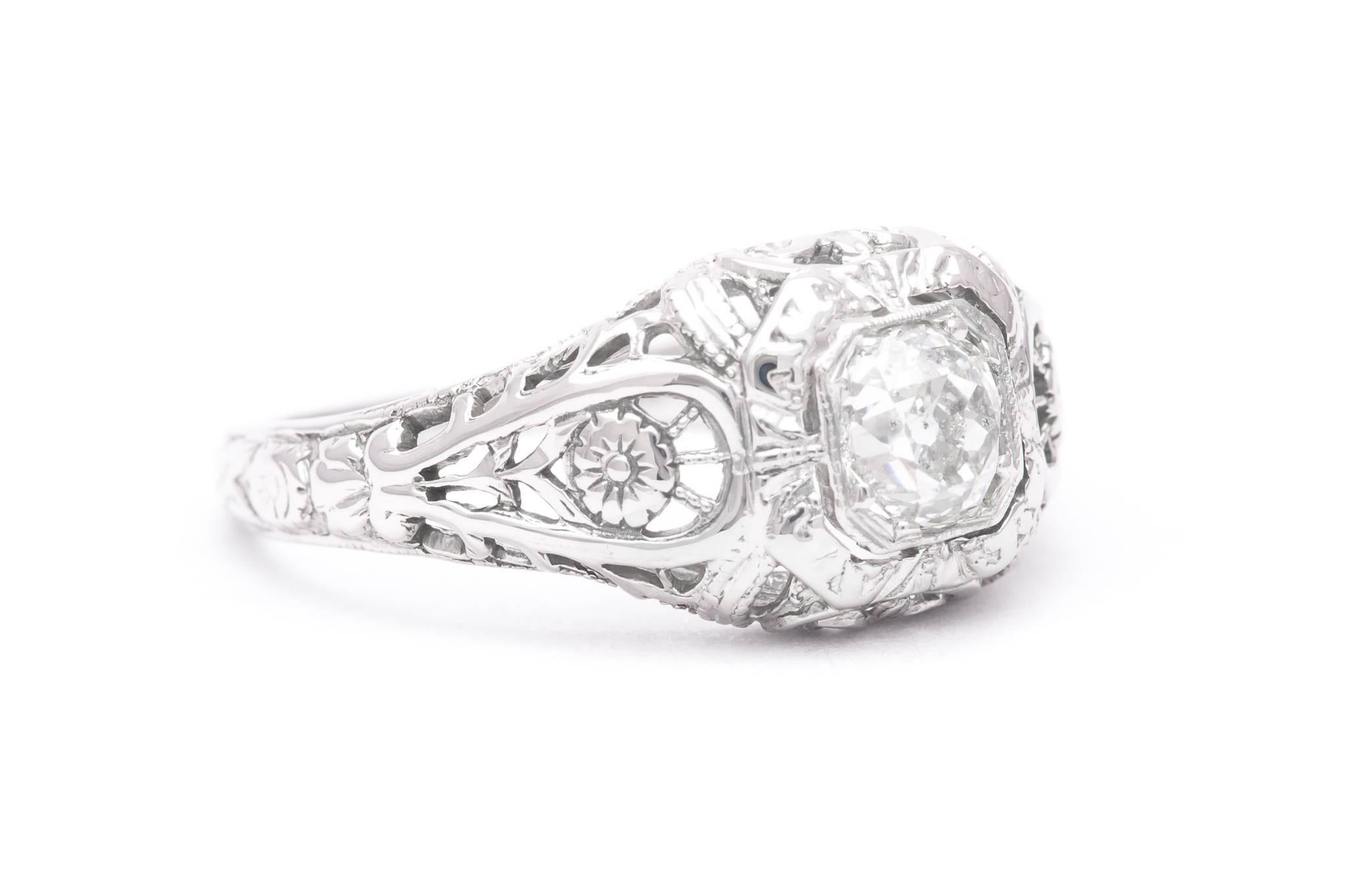 A beautiful floral motif art deco period diamond engagement ring in 18 karat white gold.  Centered by a sparkling 0.50 carat antique mine cut diamond this ring features beautiful hand pierced filigree work throughout complemented by superbly