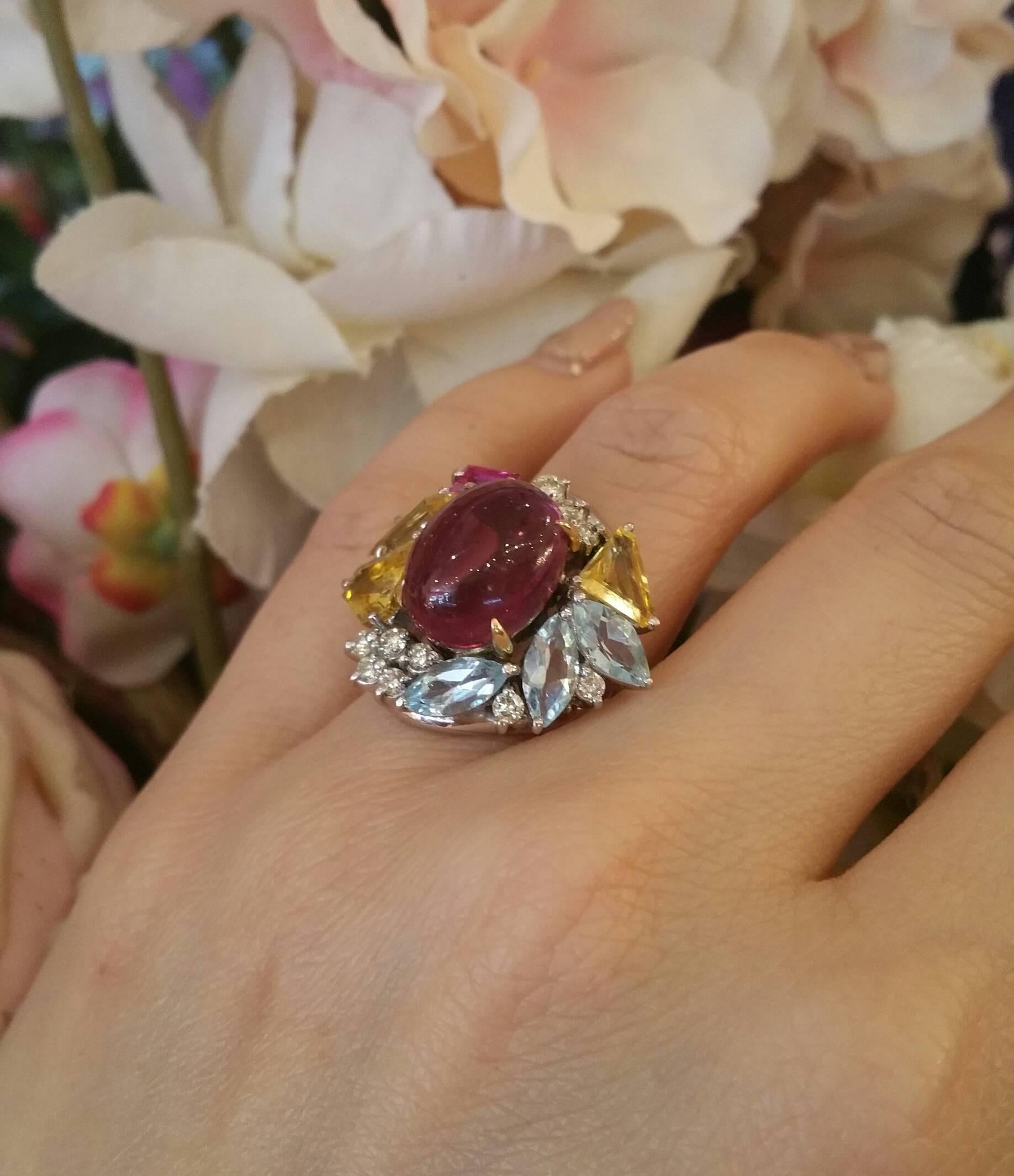 Pink Tourmaline cocktail ring featuring a 6.86 ct oval shaped cabochon in the center, surrounded by 3 fancy cut yellow Sapphires, 3 marquise shaped Aquamarines and 1 rectangular shaped Ruby, accented with round brilliant cut diamonds. (0.43ct ) 