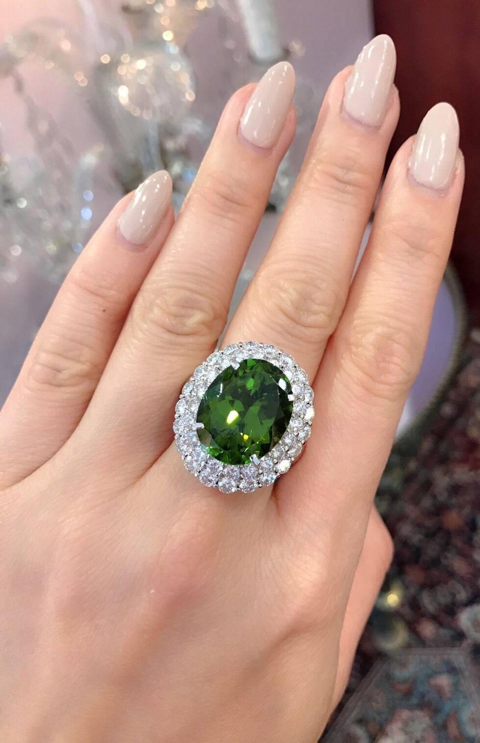 Large Oval Peridot surrounded by two rows of White and Clean Round Brilliant Diamonds in Platinum. The peridot weighs 14.67 carats, with a beautiful deep yellow-green color. It is oval shaped and faceted. The ring has two rows of Round Diamonds. The